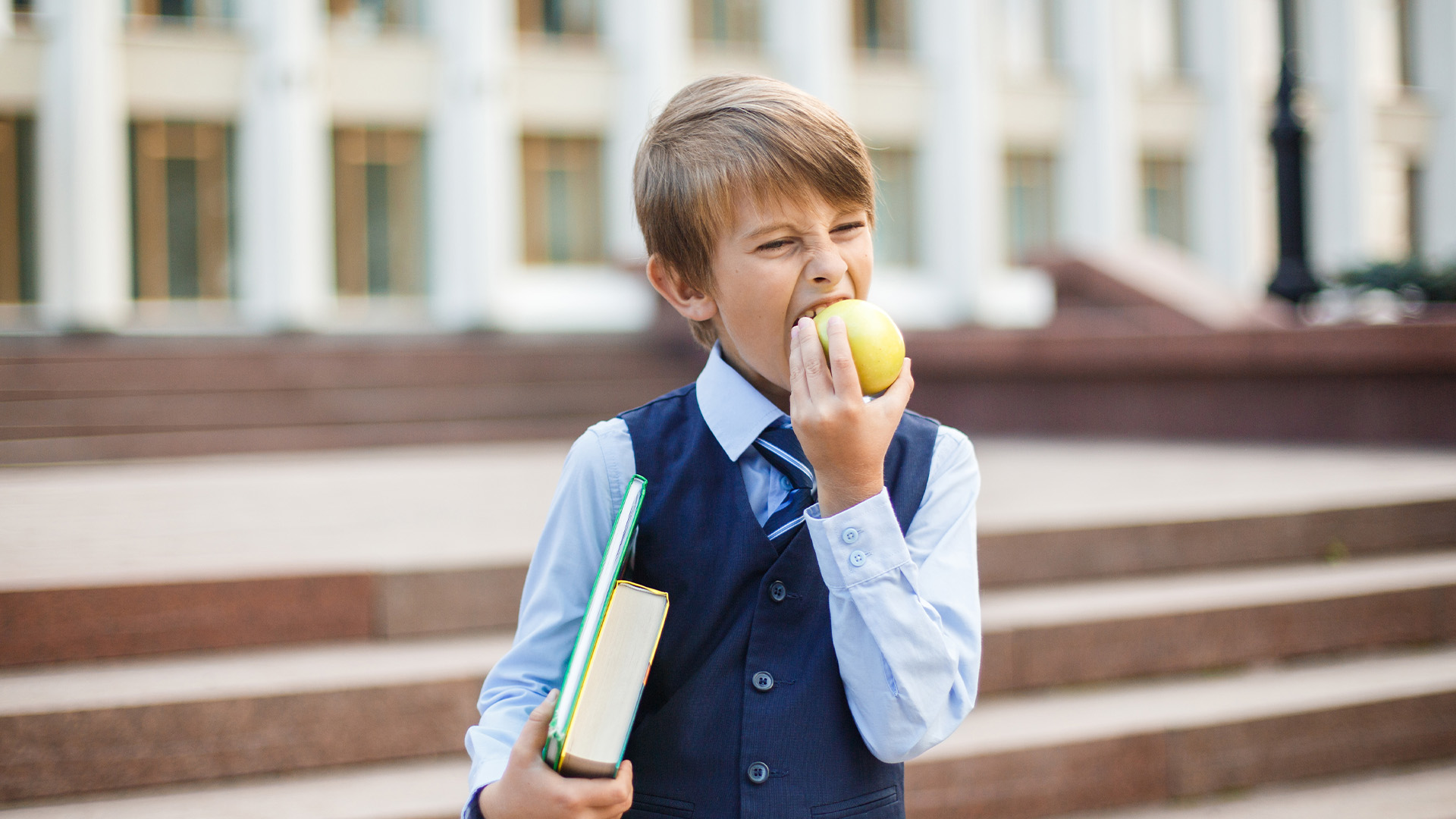 Schüler mit Schulbüchern beißt vor einem Schulgebäude in einen Apfel.