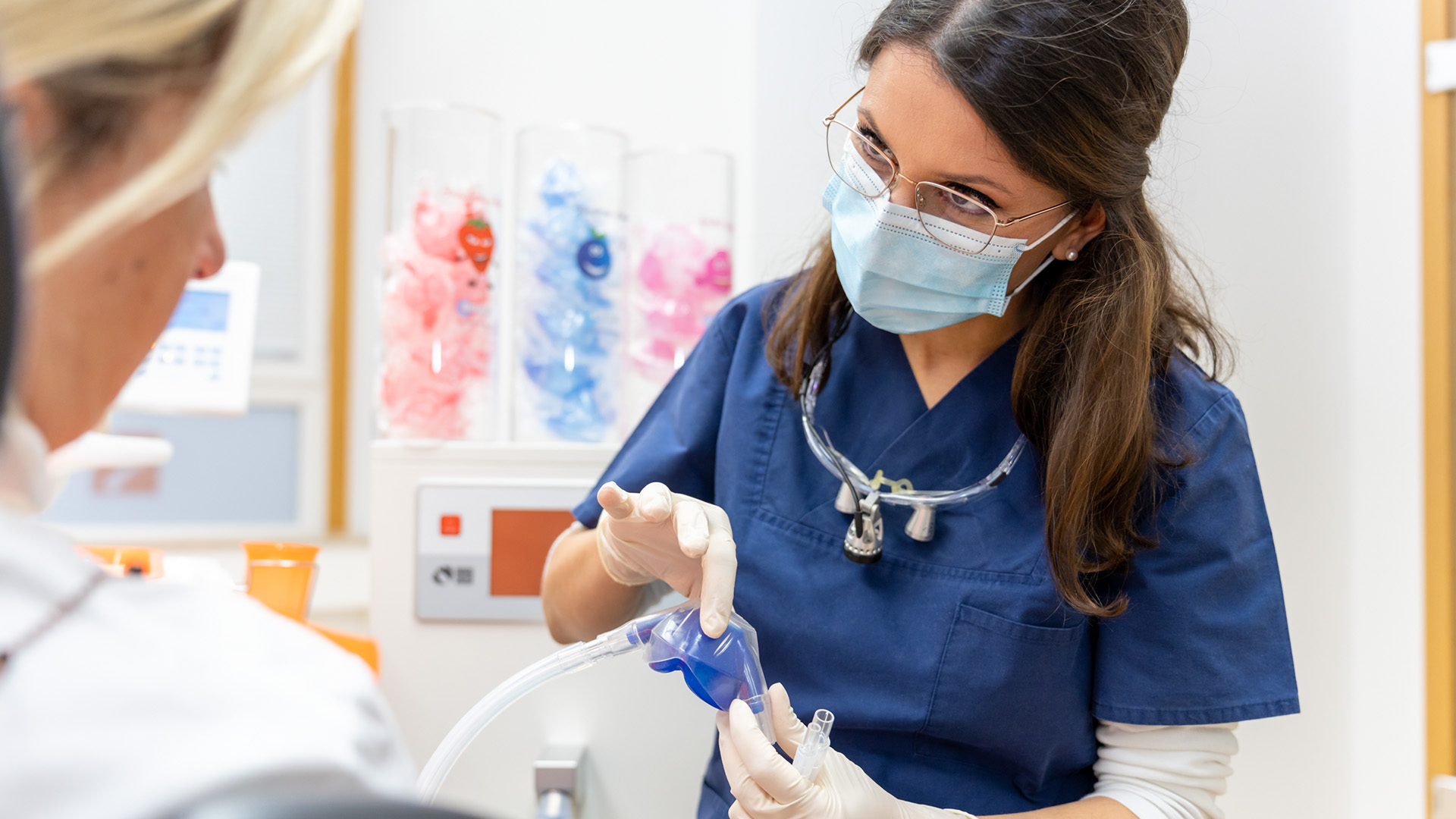 Mitarbeiterin zeigt einer Patientin eine Nasenmaske mit Schlauch.
